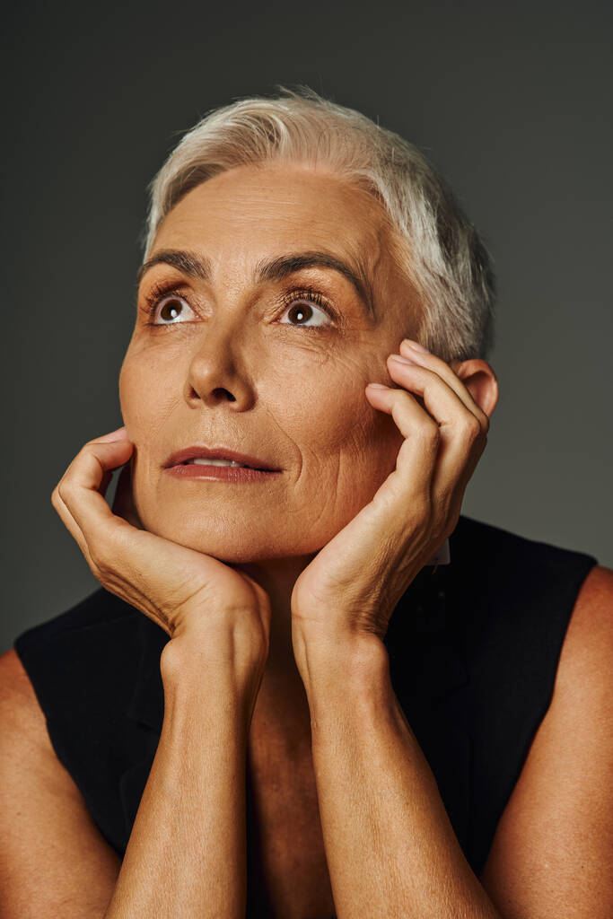 portrait de femme âgée rêveuse aux cheveux courts argentés regardant loin avec les mains près du visage sur le gris - Photo, image