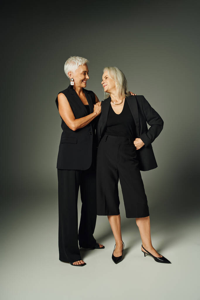 χαρούμενες ηλικιωμένες φίλες με μαύρα στυλάτα ρούχα να χαμογελούν η μία στην άλλη σε γκρι, μοντέρνα γήρανση - Φωτογραφία, εικόνα