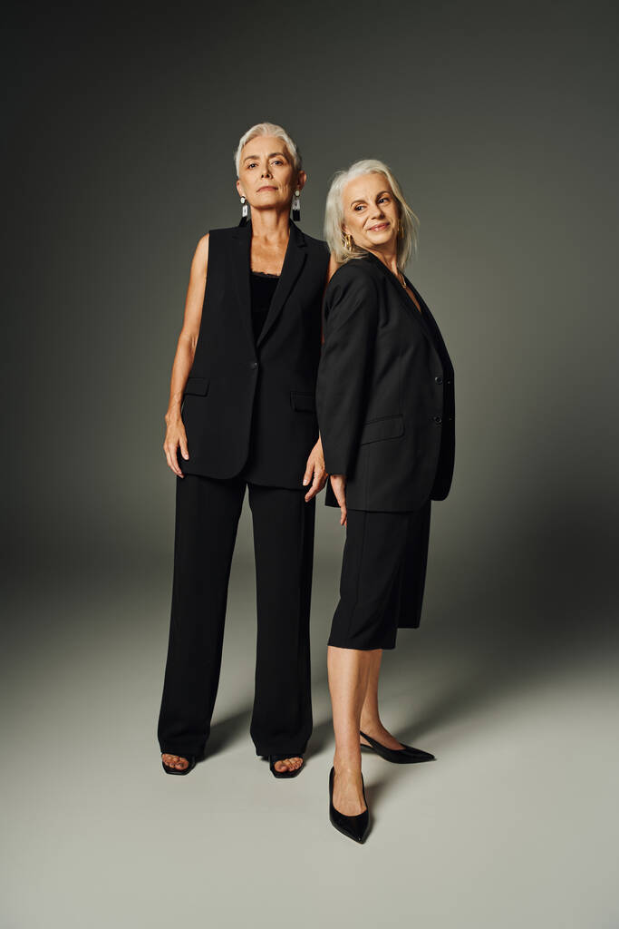 piena lunghezza di donne anziane graziose in abiti neri alla moda in piedi su grigio, moda classica - Foto, immagini