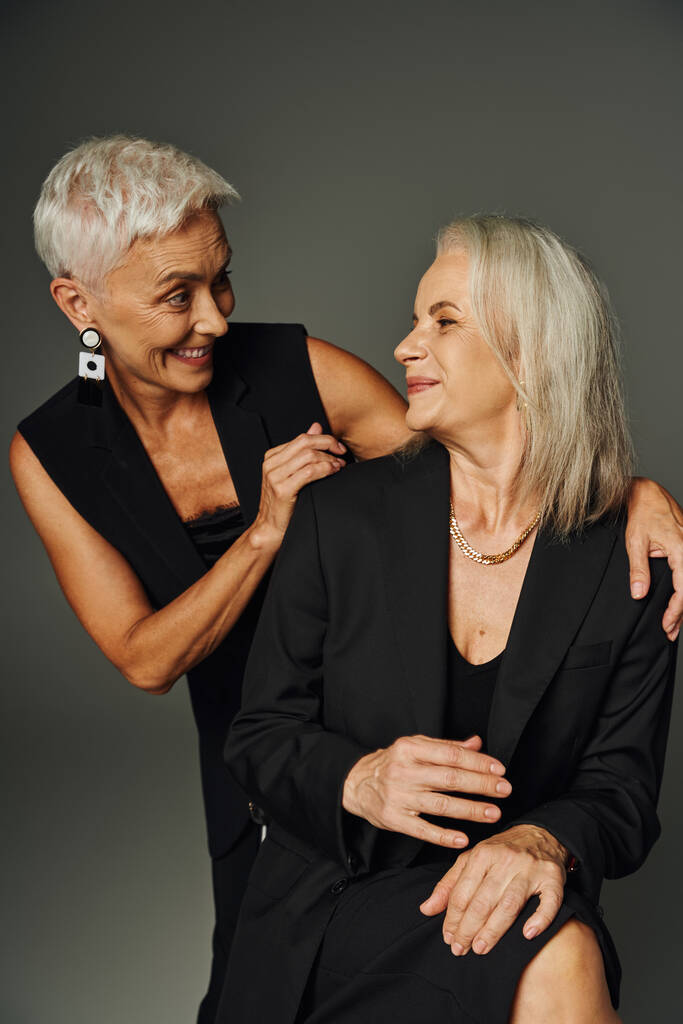 vrolijke volwassen dames in zwart modieuze kleding glimlachen naar elkaar op grijs, levenslange vriendschap - Foto, afbeelding