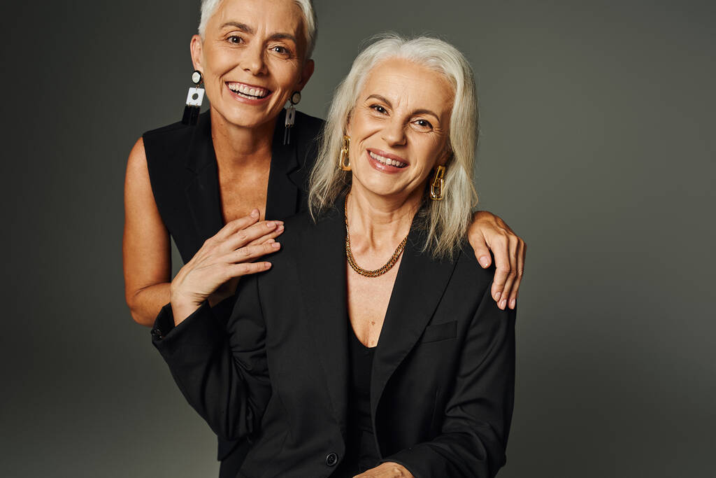 fröhliche und elegante ältere Damen in schwarzer Freizeitkleidung blicken vor grauem Hintergrund in die Kamera - Foto, Bild