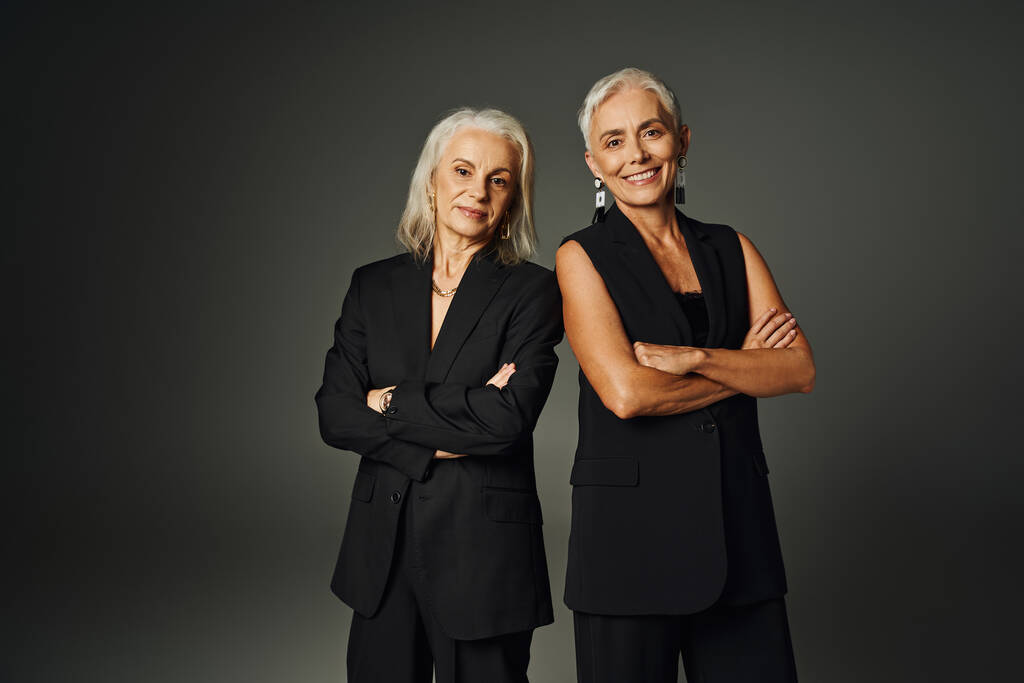 улыбающиеся старшие модели в черной элегантной одежде, стоящие со сложенными руками на серой, пожилой моде - Фото, изображение