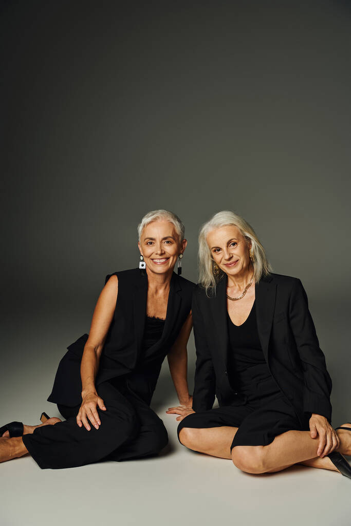 fashionable mature ladies in black clothes sitting and smiling at camera on grey, classic fashion - Photo, Image