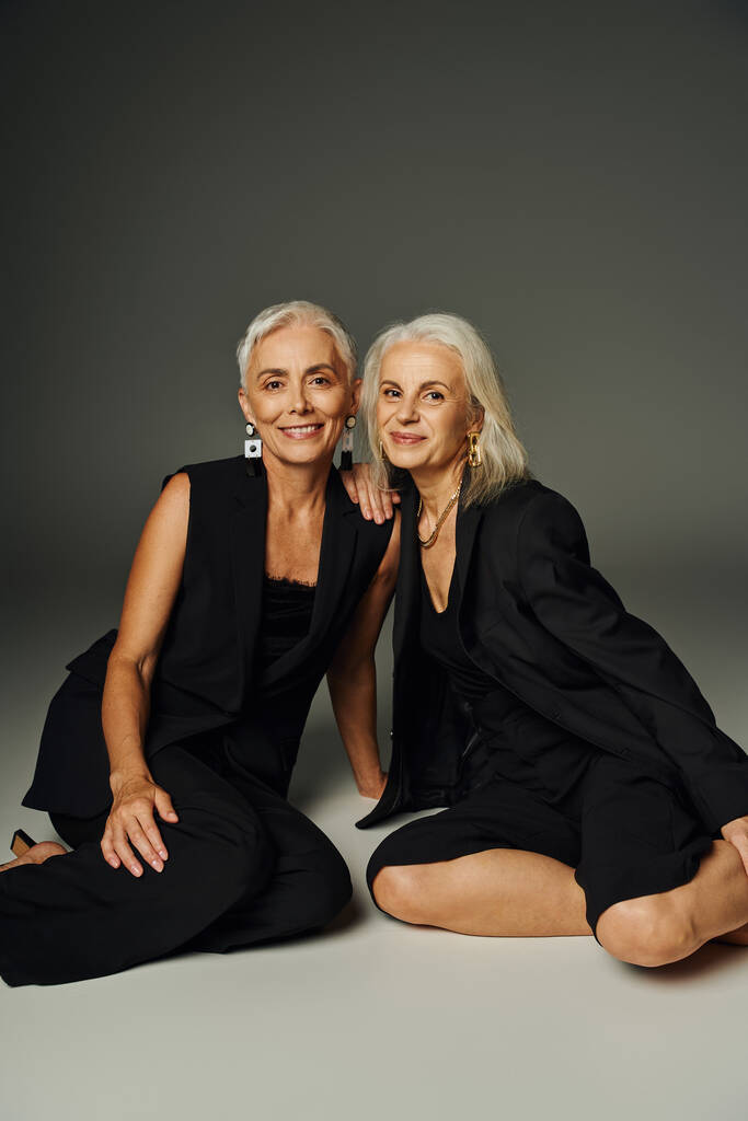 modelos senior sonrientes en traje elegante negro sentado sobre fondo gris, envejecimiento elegante y amistad - Foto, Imagen