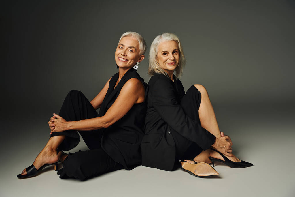χαρούμενες και μοντέρνες ηλικιωμένες γυναίκες με μαύρα ρούχα που κάθονται πλάτη με πλάτη σε γκρι φόντο, κομψότητα - Φωτογραφία, εικόνα