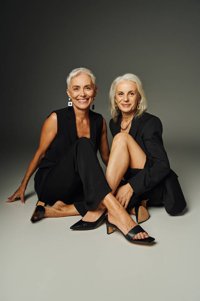 Positive Senior-Trendsetter in schwarzer Kleidung sitzen auf grauer, klassischer Mode und lächeln in die Kamera - Foto, Bild