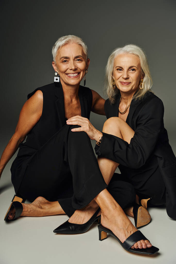 очаровательные элегантные старшие модели в черной одежде сидят и улыбаются в камеру на сером, стильное старение - Фото, изображение
