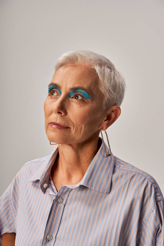 élégant modèle senior rêveur avec cheveux courts argentés et eye-liner bleu regardant loin sur gris, portrait - Photo, image
