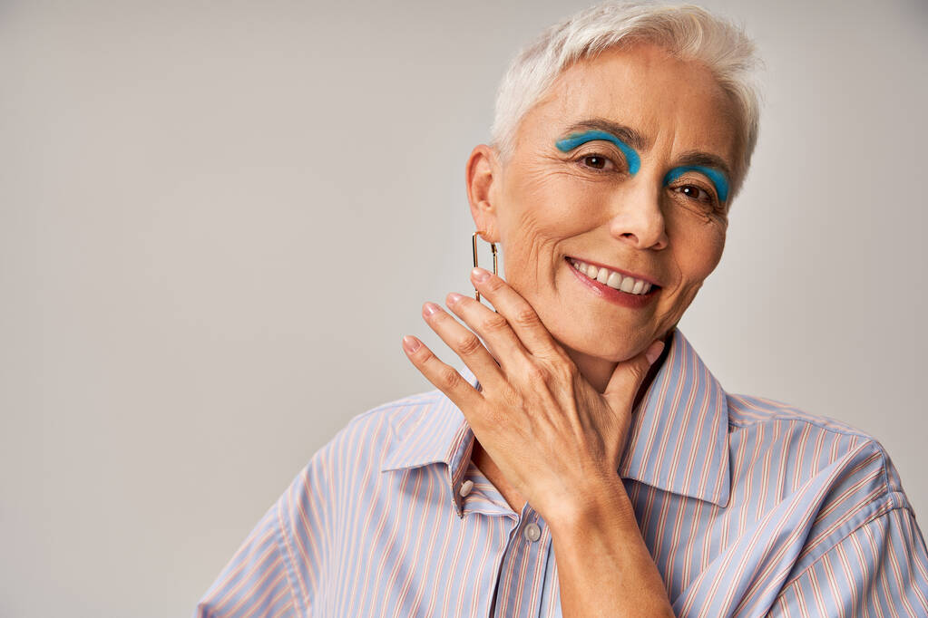 cheerful mature lady with blue eyeliner and short silver hair looking at camera on grey backdrop - Photo, Image