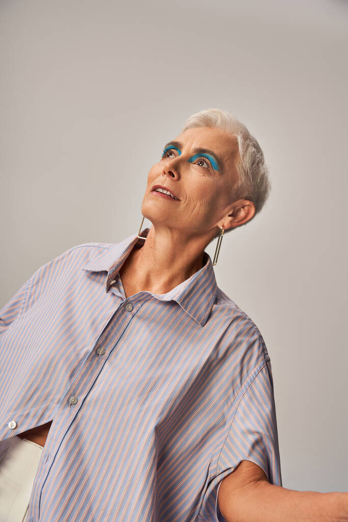 donna anziana sognante e alla moda con trucco audace e capelli corti d'argento distogliendo lo sguardo sul grigio - Foto, immagini