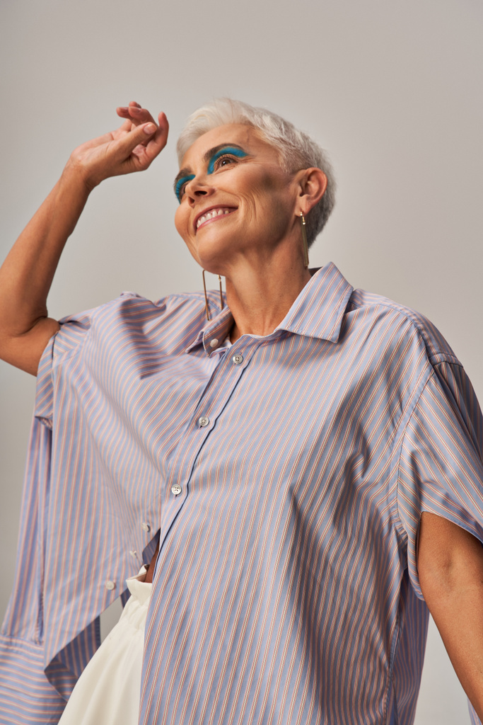 χαρούμενη και μοντέρνα ηλικιωμένη γυναίκα με τολμηρό μακιγιάζ και κοντά ασημένια μαλλιά κοιτάζοντας μακριά σε γκρι φόντο - Φωτογραφία, εικόνα