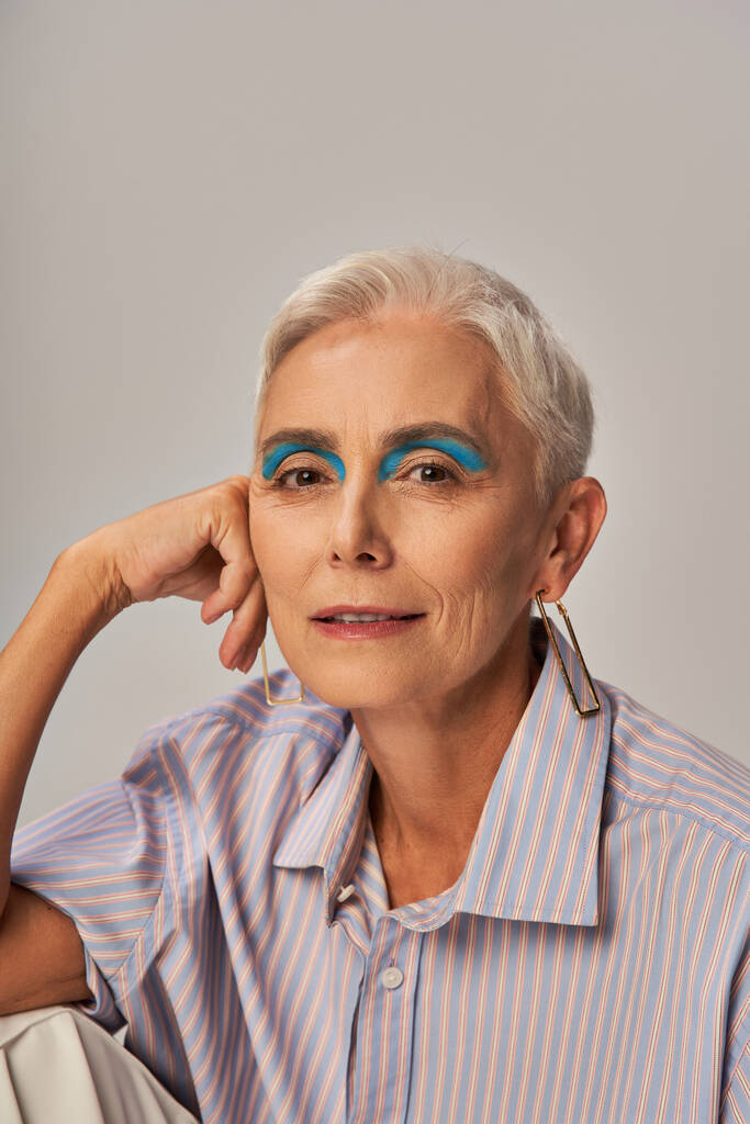 trendy senior model met kort zilverkleurig haar en blauwe eyeliner kijkend naar camera op grijs, portret - Foto, afbeelding