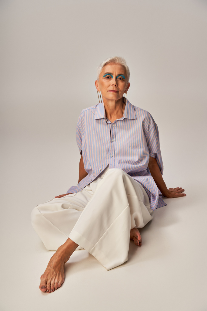 босоніж і впевнена старша леді в модному повсякденному вбранні дивиться на камеру, сидячи на сірому - Фото, зображення