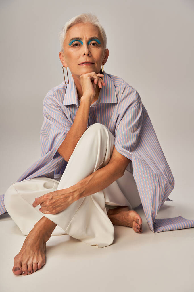 μοντέρνα γήρανση, ξυπόλυτη ώριμη γυναίκα σε μπλε ριγέ πουκάμισο κάθεται και κοιτάζοντας κάμερα σε γκρι - Φωτογραφία, εικόνα