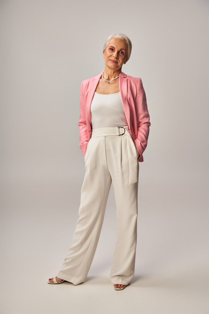 volledige lengte van senior model in roze blazer staande met de handen in zakken van witte broek op grijs - Foto, afbeelding