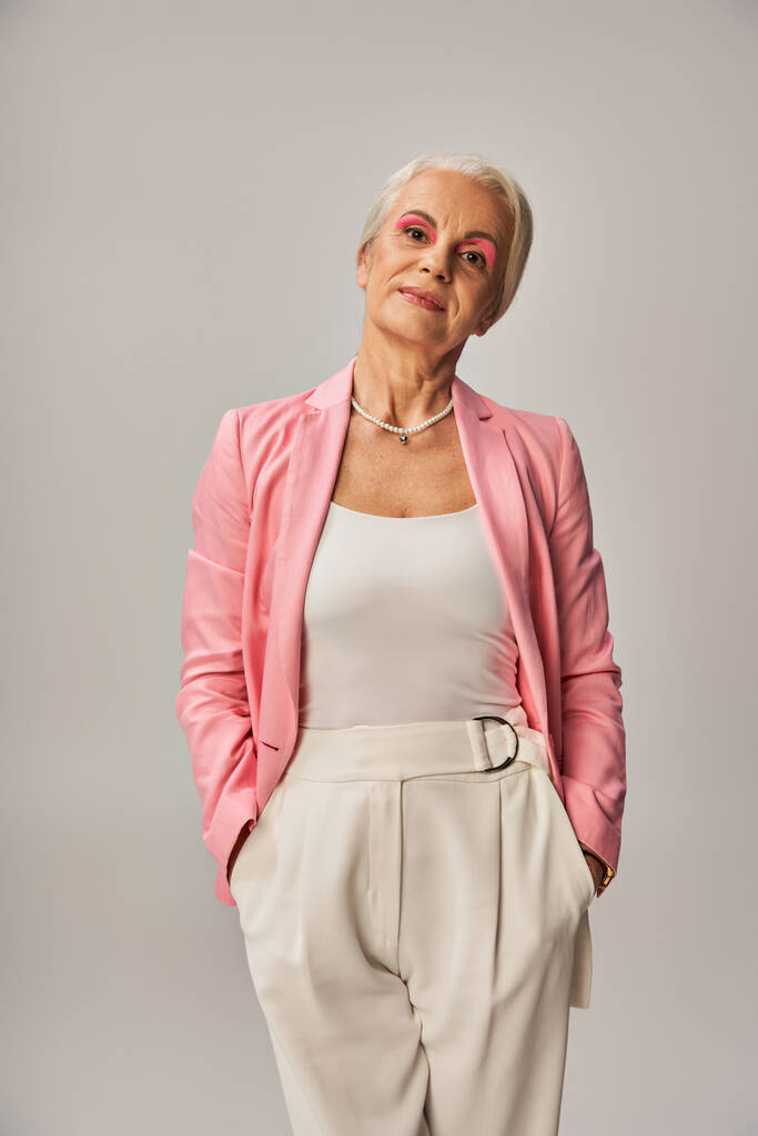 trendige reife Dame im rosafarbenen Blazer stehend mit den Händen in Taschen weißer Hosen auf grau, Eleganz - Foto, Bild
