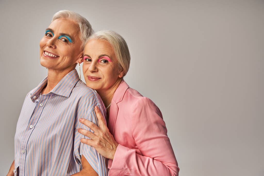 enchanté les amies aînées en vêtements bleus et roses souriant à la caméra sur gris, âge positivité - Photo, image