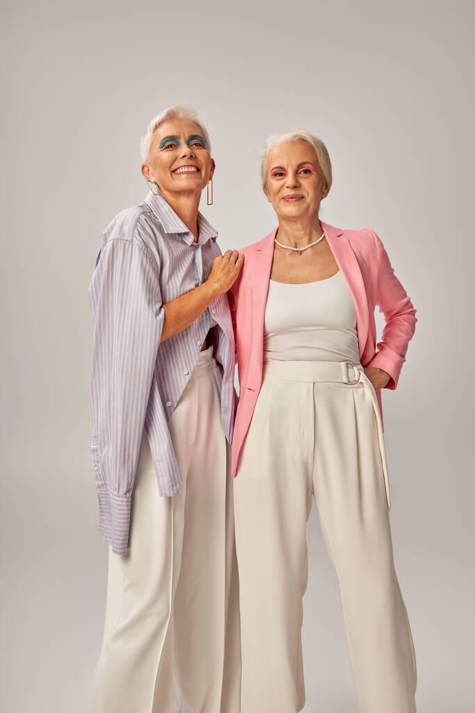 alegres señoras mayores en atuendo casual de moda mirando a la cámara en gris, concepto de envejecimiento feliz - Foto, imagen