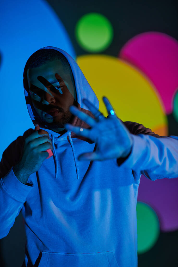 good looking man with hood on putting hand in front of camera in projector lights, fashion concept - Photo, Image