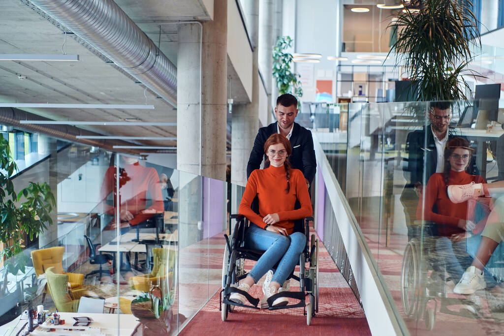 Yrityksen johtaja avustaa liikekumppaniaan pyörätuolissa, auttaa häntä navigoimaan startup-toimistoon, jossa he työskentelevät yhdessä monipuolisen kollegojensa kanssa korostaen osallisuutta ja - Valokuva, kuva