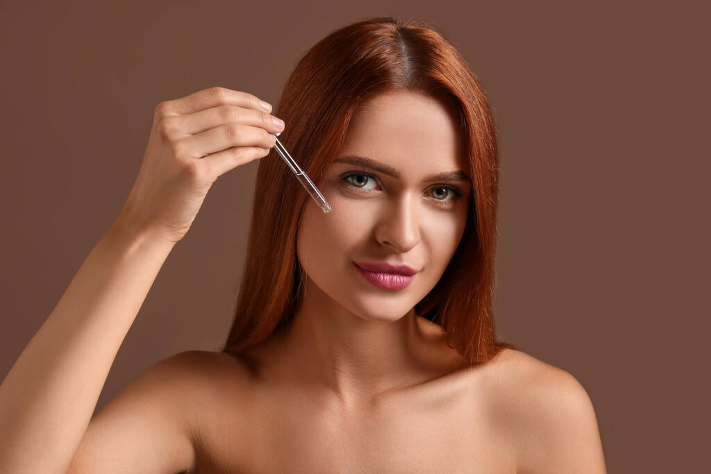 Piękna młoda kobieta nakładająca serum kosmetyczne na twarz na brązowym tle - Zdjęcie, obraz