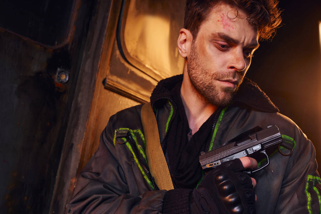 hombre sin afeitar y sombrío con la cara arañada mirando el arma en el metro oscuro, refugio después del desastre - Foto, Imagen