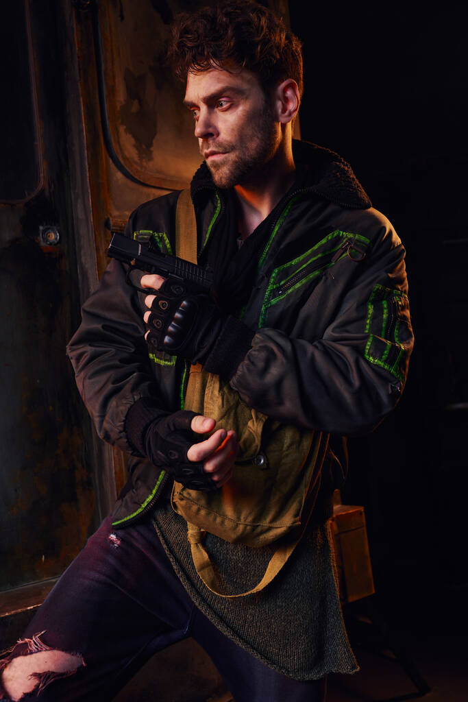 Ongeschoren man in versleten jasje en vingerloze handschoenen met pistool in donkere metro, post-apocalyps - Foto, afbeelding