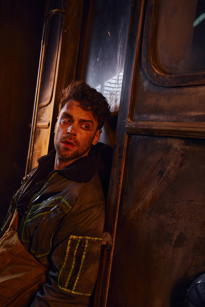 neoholený muž v bundě odvrací pohled v blízkosti rezavého kočáru v nebezpečném prostředí opuštěného metra - Fotografie, Obrázek