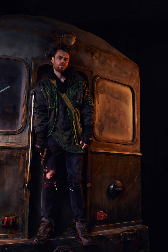 оставшийся в живых после апокалипсиса в изношенной одежде, стоящий с пистолетом на ржавом вагоне метро в подполье - Фото, изображение