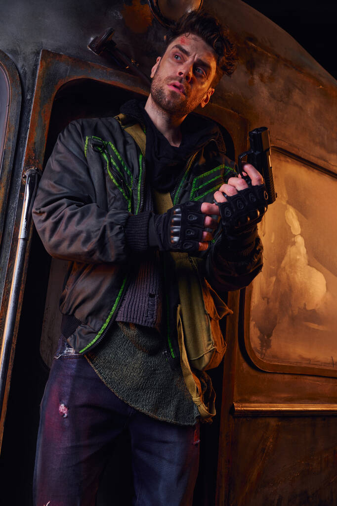 nieogolony mężczyzna w zużytych ubraniach i rękawiczkach bez palców trzymający broń i odwracający wzrok w opuszczonym metrze - Zdjęcie, obraz