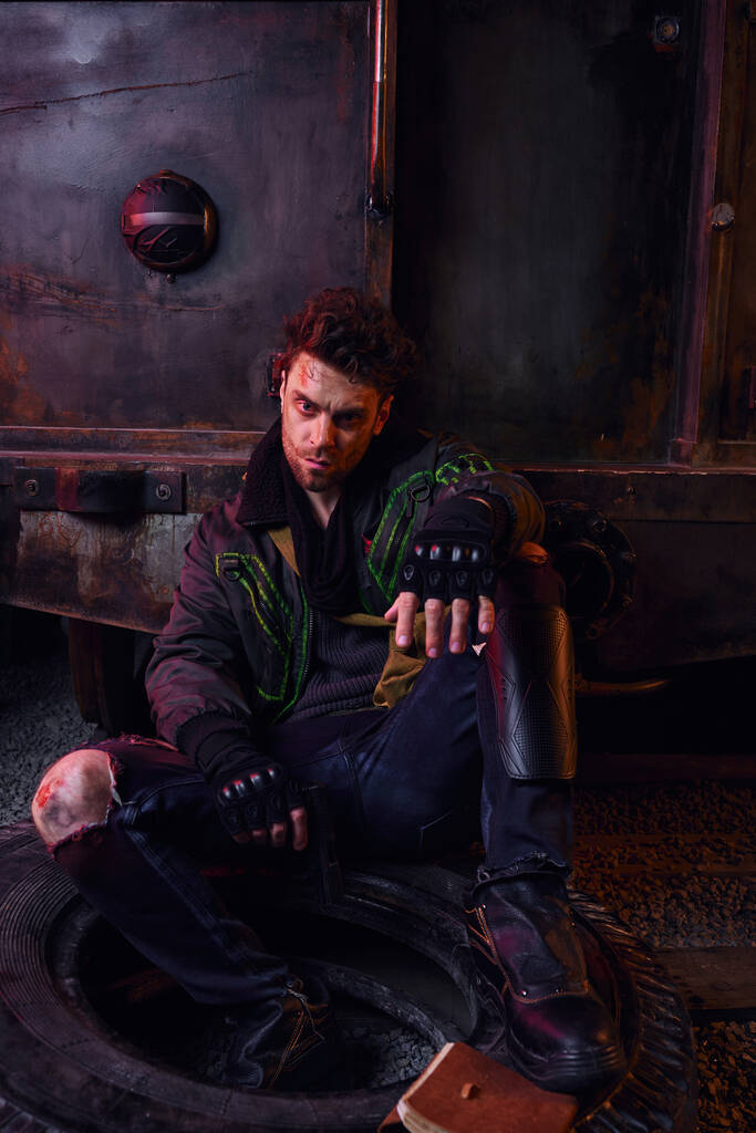 Похмурий чоловік у зношеному вбранні, сидячи на шині з пістолетом у покинутому метро, життя після ядерної катастрофи - Фото, зображення