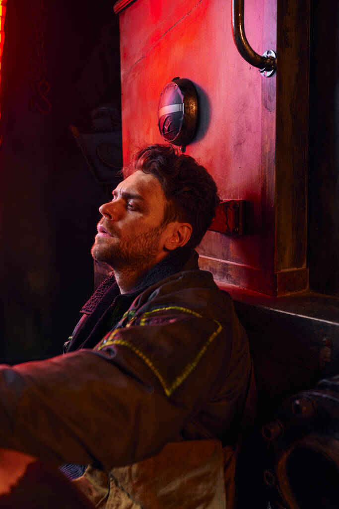 vista lateral del hombre deprimido sin afeitar sentado cerca de un vagón oxidado en el metro, desesperación post-apocalíptica - Foto, imagen