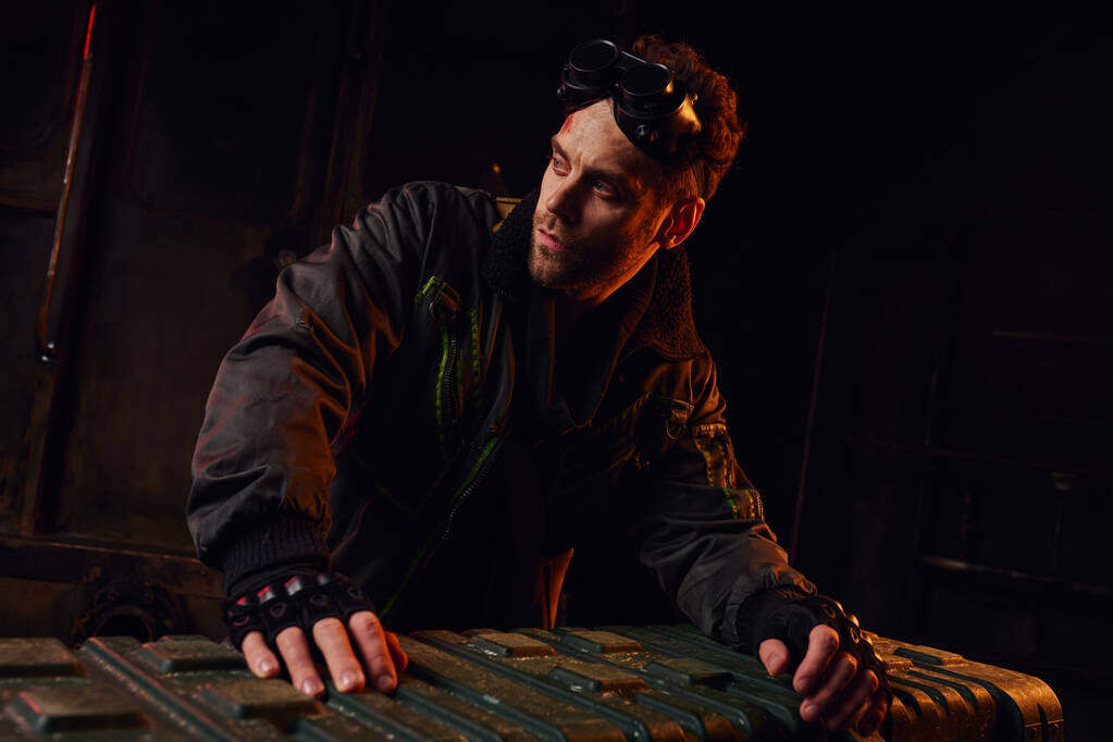 homme non rasé avec googles regardant loin près de la boîte à armes dans l'obscurité post-apocalyptique souterraine - Photo, image