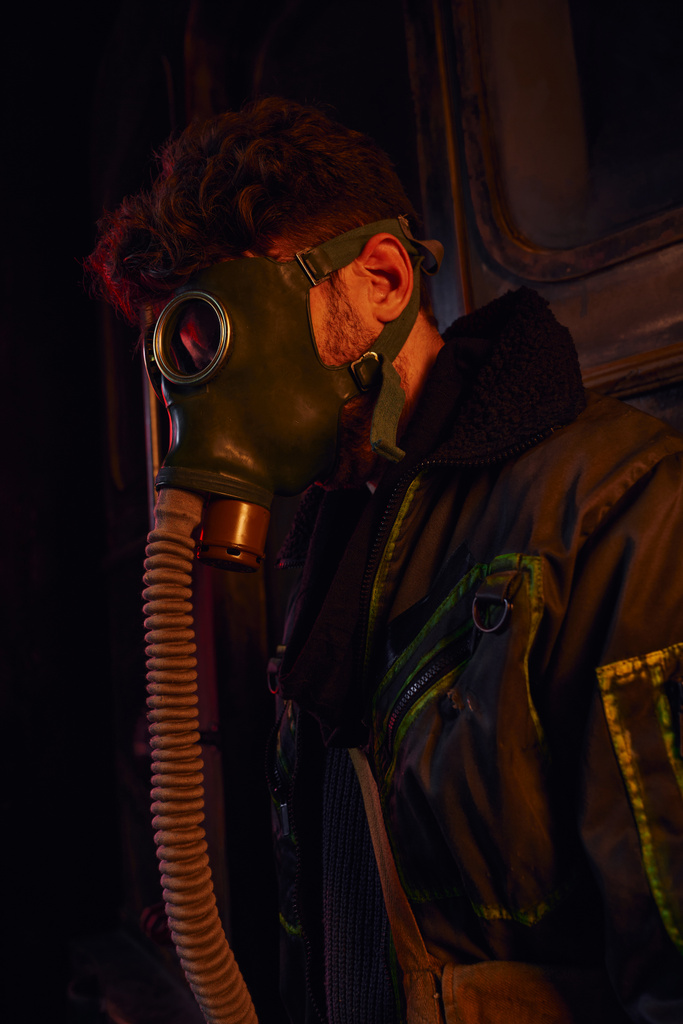 Maskeli ve ceketli bir adam karanlık yeraltı tünelinin kırmızı ışığı altında, kıyamet sonrası hayatta kalma mücadelesi veriyor. - Fotoğraf, Görsel