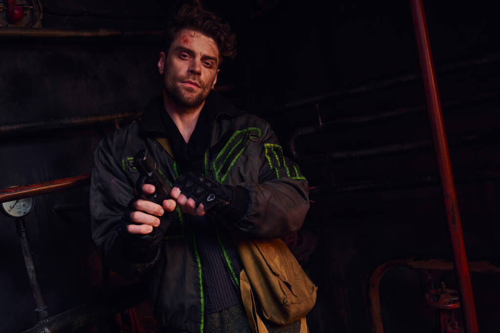 sebevědomý muž s poškrábaným obličejem nabíjení zbraň a při pohledu na kameru v tmavém podzemním tunelu - Fotografie, Obrázek
