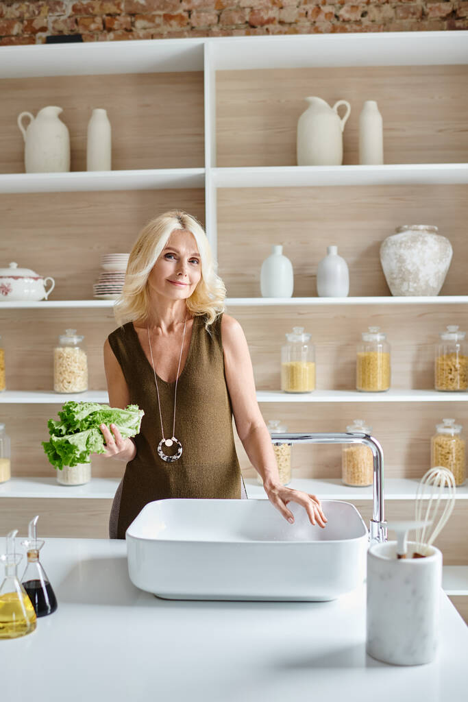 atractiva mujer vegetariana de mediana edad con cabello rubio lavando lechuga fresca en fregadero de cocina - Foto, imagen
