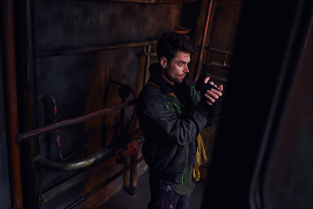 Ongeschoren man met pistool die wegkijkt in donkere ondergrondse tunnel bij roestige buizen, post-apocalyps - Foto, afbeelding