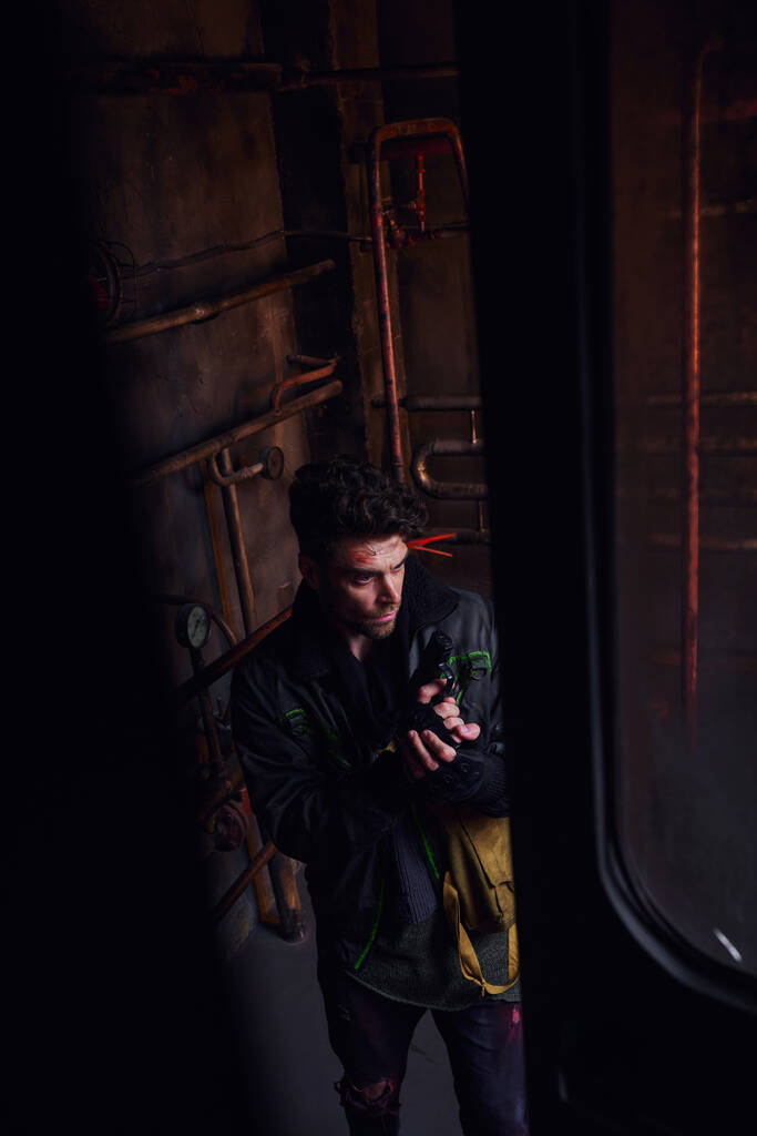 vysoký úhel pohledu na muže v ošoupaných šatech stojící s pistolí v postapokalyptickém podzemním tunelu - Fotografie, Obrázek