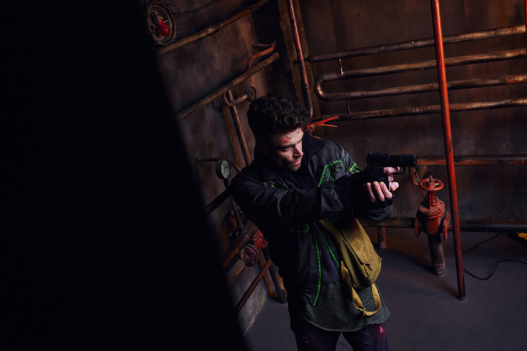 soustředěný muž mířící s pistolí v opuštěném metru v blízkosti rezavých potrubí, herní charakter - Fotografie, Obrázek