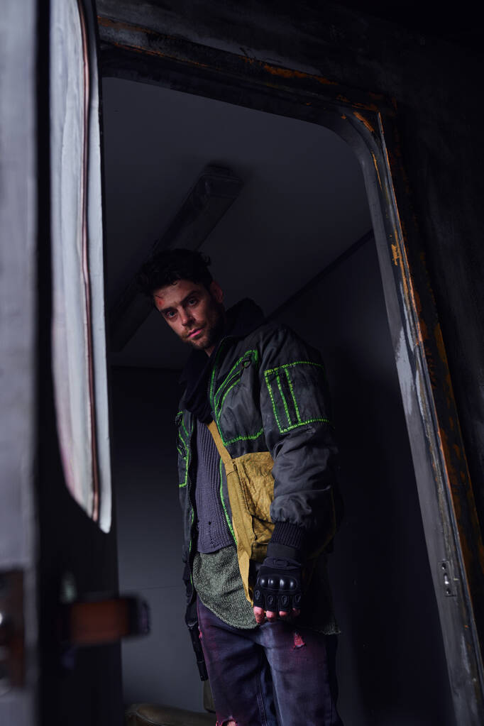 мрачный человек в черной куртке, смотрящий в камеру в заброшенном вагоне метро, постапокалиптическая концепция - Фото, изображение