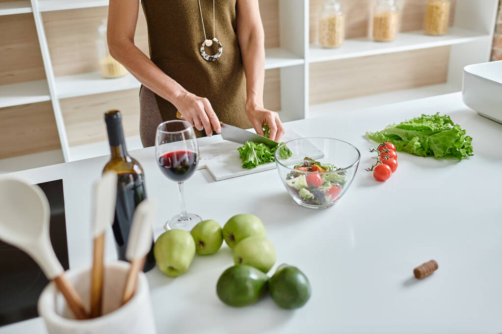 Teilaufnahme einer Frau, die frischen Salat schneidet und Gemüsesalat in der Nähe eines Rotweinglases zubereitet - Foto, Bild