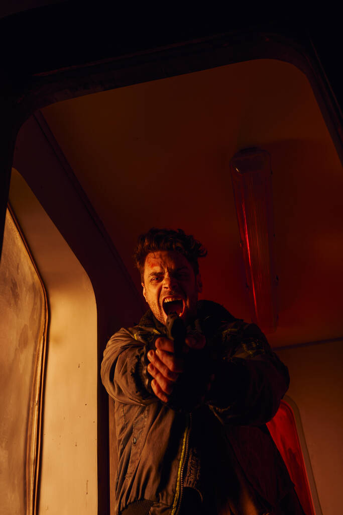 hombre gritando mirando a la cámara y apuntando con el arma en la luz roja del vagón abandonado del metro - Foto, imagen