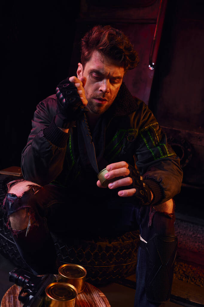 голодний чоловік у зношеному вбранні, що відкриває консервовану їжу з військовим ножем у темній посткатастрофі під землею - Фото, зображення