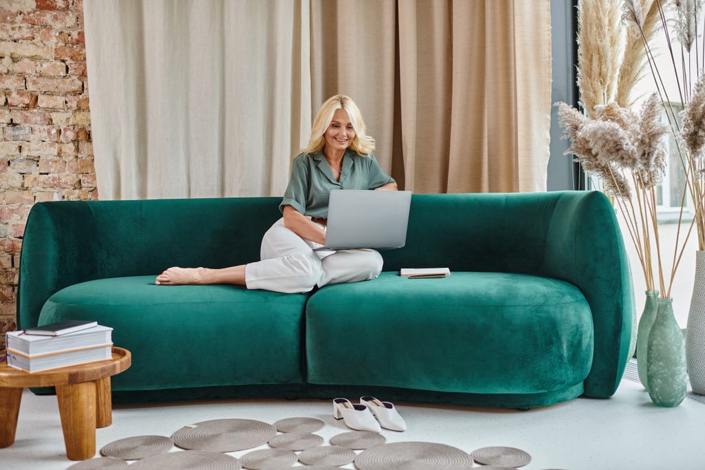 zrelaksowana kobieta w średnim wieku z blond włosami przy użyciu laptopa podczas siedzenia na kanapie, praca w domu - Zdjęcie, obraz