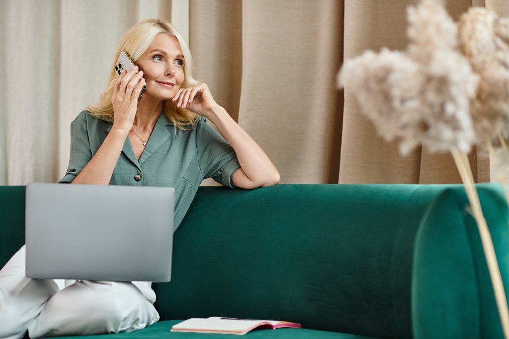 attractive middle aged woman with blonde hair talking on smartphone and using laptop on sofa - Photo, Image
