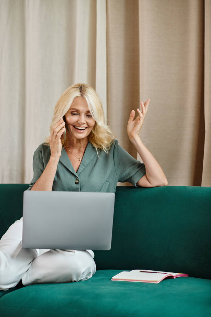веселая женщина средних лет со светлыми волосами, разговаривающая на смартфоне и пользующаяся ноутбуком на диване - Фото, изображение