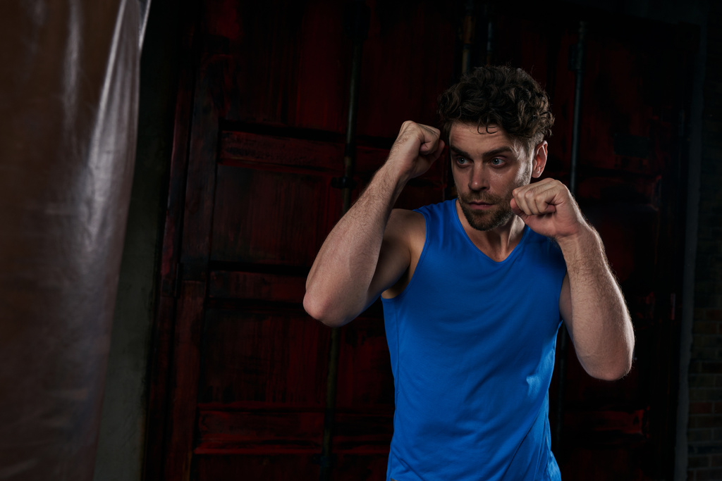 starker Mann in blauem Tanktop, der nachts in der Stadt mit Boxsack trainiert, Straßensport - Foto, Bild