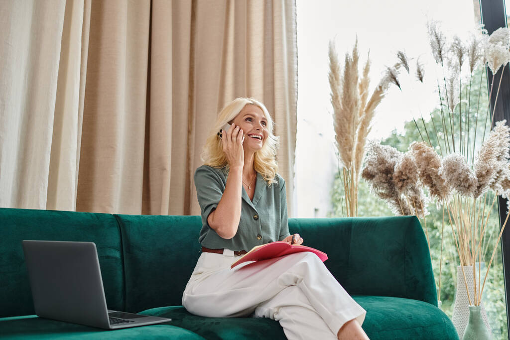 счастливая женщина средних лет разговаривает на смартфоне и делает заметки, сидя на диване с ноутбуком - Фото, изображение