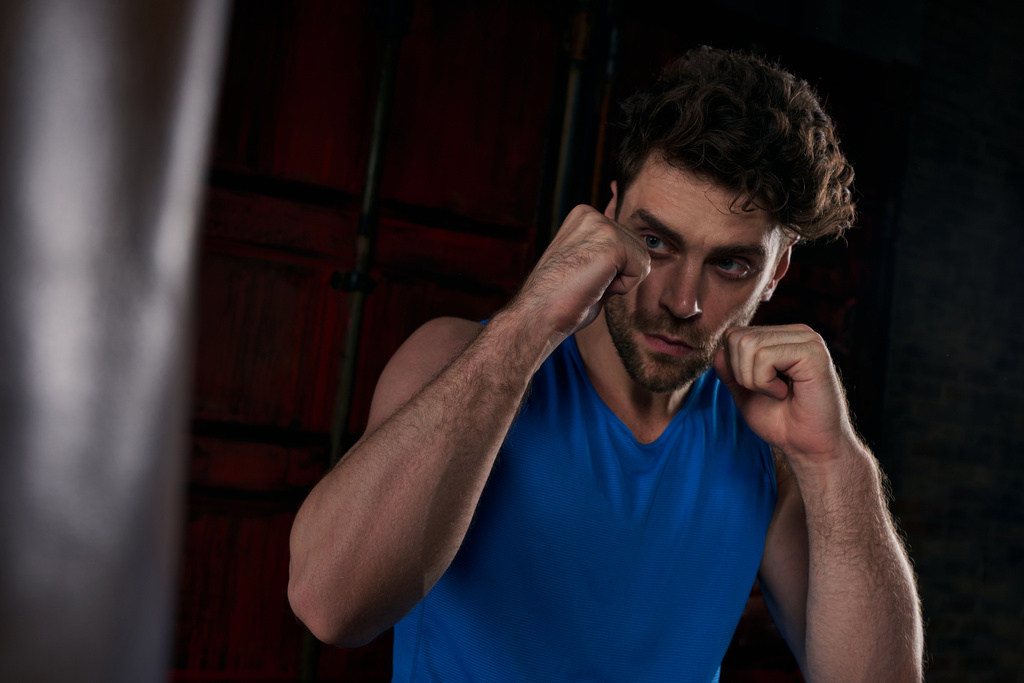 athletischer und unrasierter Mann, der in dunkler Stadt mit Boxsack trainiert, Straßensportler - Foto, Bild