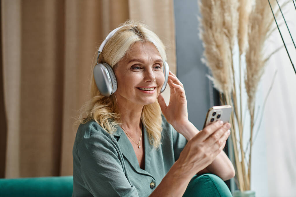 χαρούμενη μεσήλικη γυναίκα με ασύρματα ακουστικά που ακούει μουσική και χρησιμοποιεί smartphone στο σπίτι - Φωτογραφία, εικόνα
